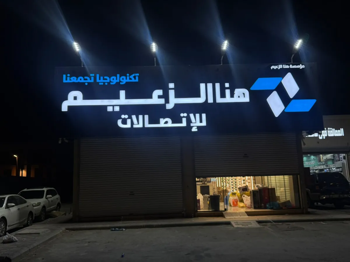 لوحة هنا الزعيم للاتصالات مضيئة ليلاً بأحرف بيضاء وشعار أزرق