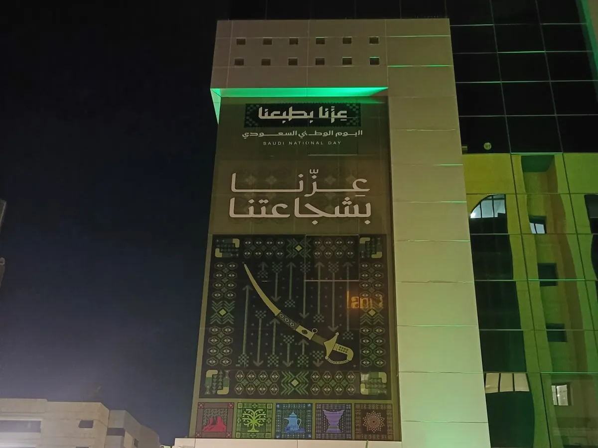 تغليف عمود واجهة مبنى بتصميم اليوم الوطني «عزنا بطبعنا» مع إضاءة خضراء ليلاً