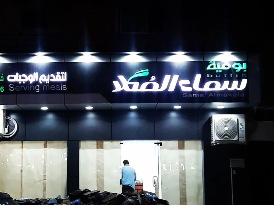 لوحة بوفية سماء المكلا بأحرف بارزة مضيئة وكشافات علوية على واجهة سوداء ليلاً